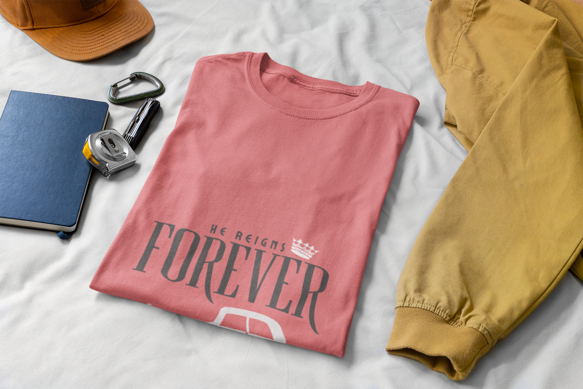 folded Pink t-shirt with 'We Reigns Forever' text on a white surface with yellow pants and accessories.