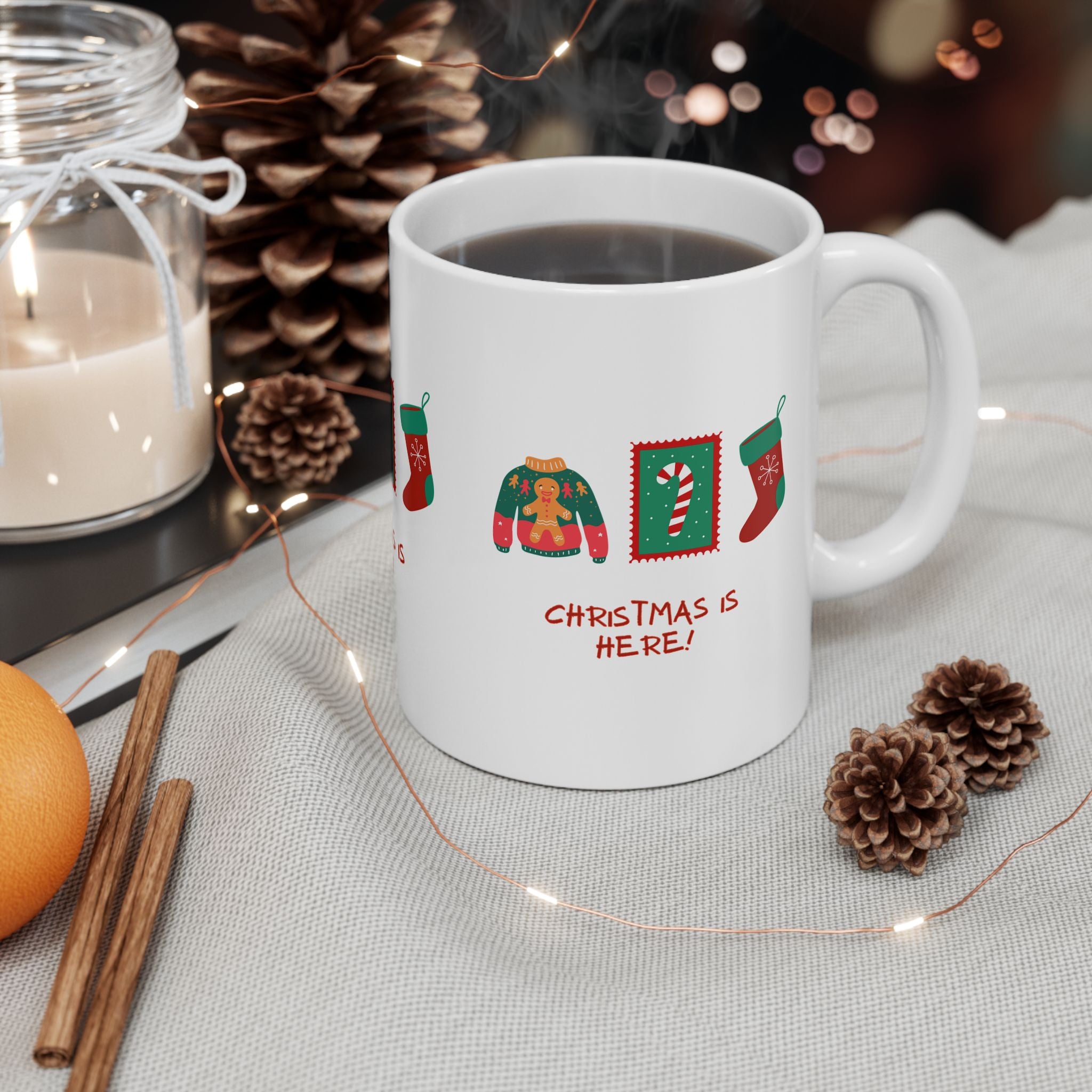 White mug with Christmas-themed design and text 'Christmas is here!' on a table with decorative items.