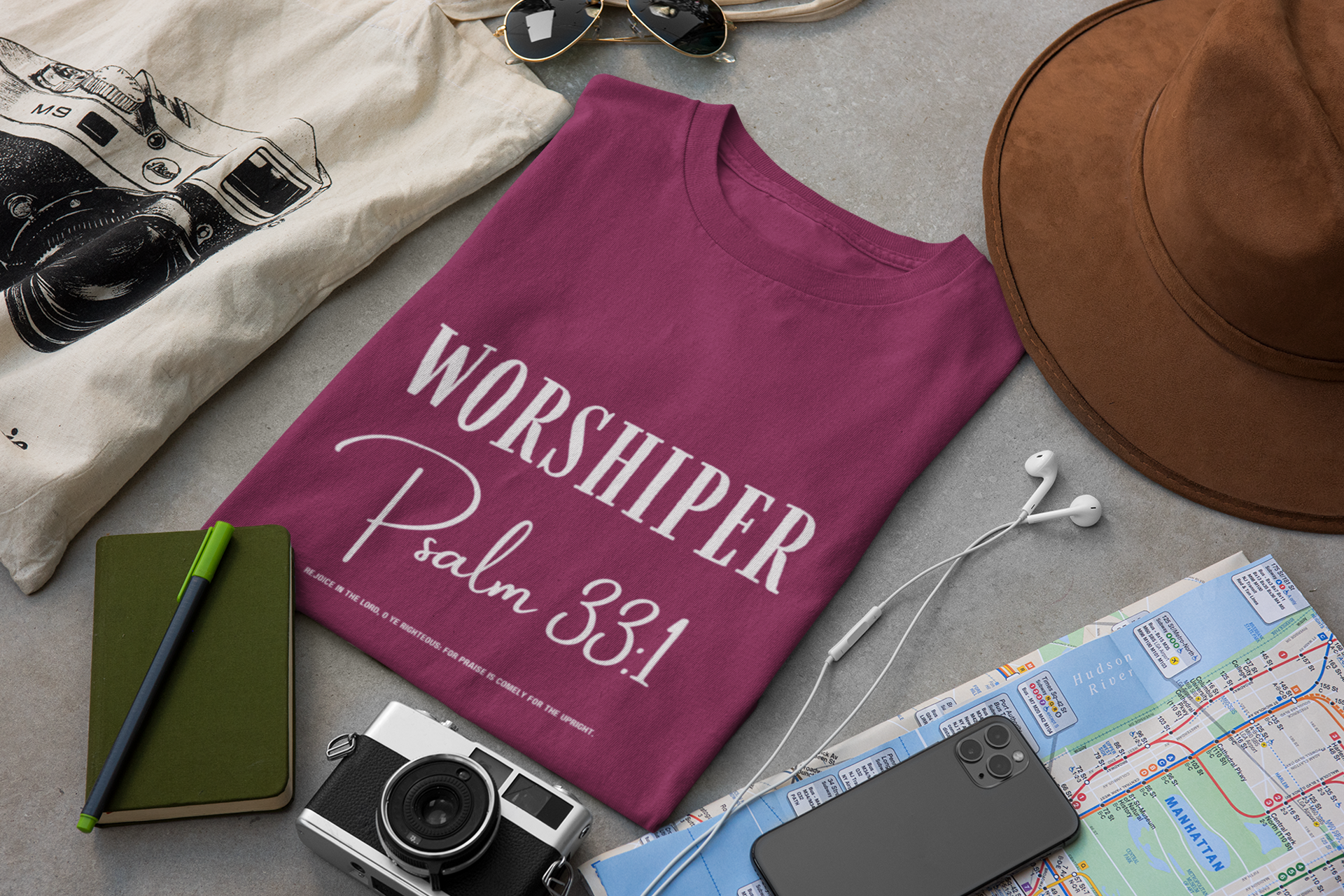 folded Purple t-shirt with 'Worshiper Psalm 33:1' text on table with hat, map, phone, sunglasses
